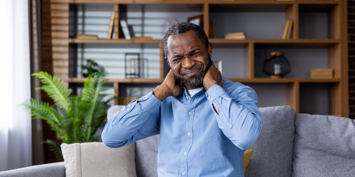 Man sitting on sofa in living room experiencing neck pain. Comfortable home setting with potted plant and bookshelf in background. Concept of discomfort, pain relief, home interior.