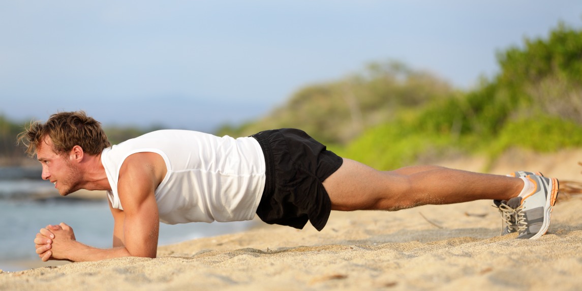 Crossfit training fitness man doing plank core exercise working out his midsection core muscles. Fit male fitness instructor planking exercising outside in summer on beach.