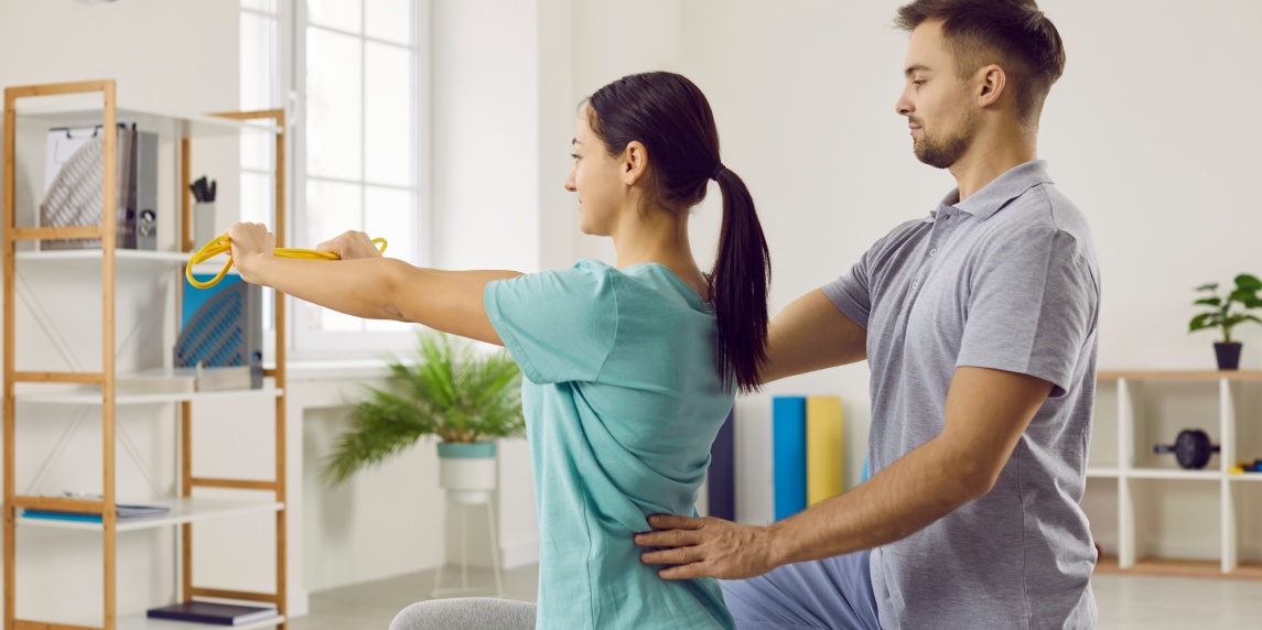 Physical recovery with rehabilitation specialist. Female patient with elastic band does stretching exercise with male physiotherapist. Doctor helps girl to correct wrong posture after back injury