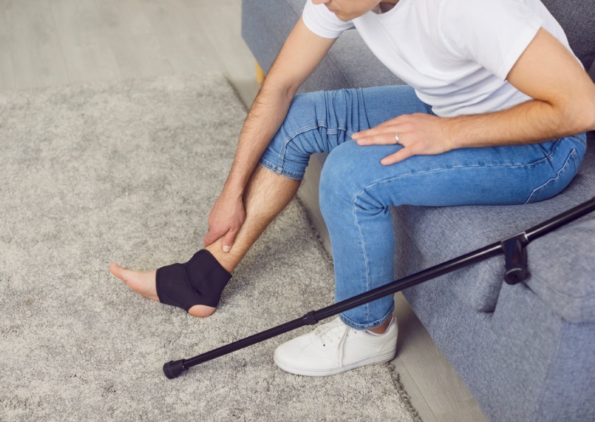 Young man with crutches in casual clothes massaging his injured leg sitting on sofa at home in black bandage. Top view of male person with sprain after accident. Rehabilitation and injury concept.