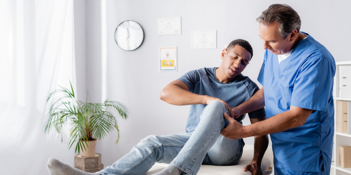 mature chiropractor touching injured knee of african american patient