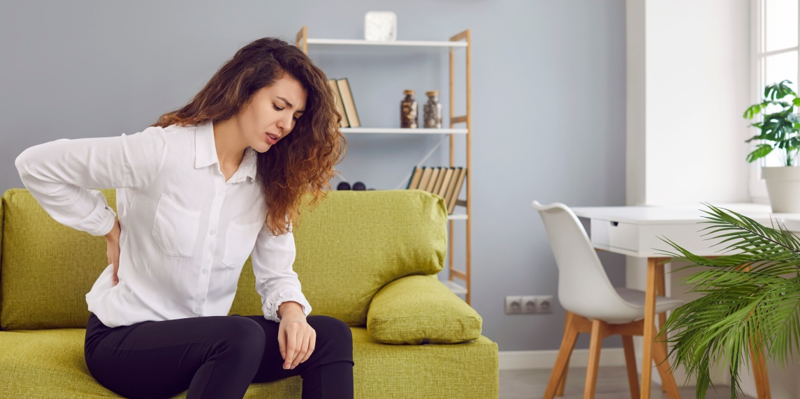 Woman suffering from strong lumbago pain. Beautiful young lady with chronic backache is sitting on the sofa at home, feeling pain, and holding her hand on her lower back