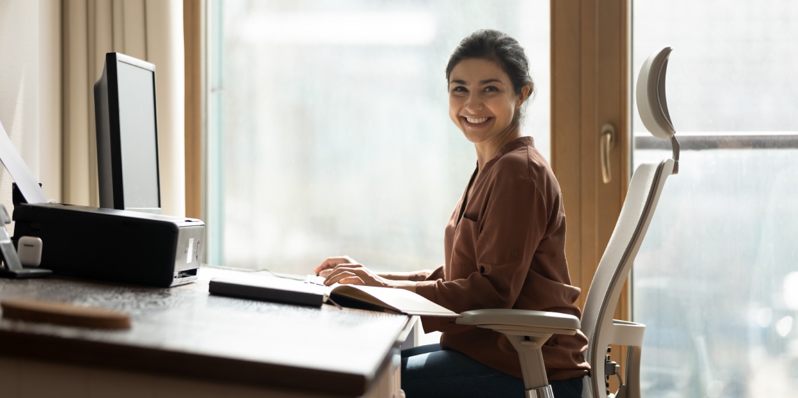 Modern day worker. Portrait of happy biracial business woman freelancer sit by computer at comfy workplace at corporate workspace or at home. Smiling young indian lady office employee look at camera